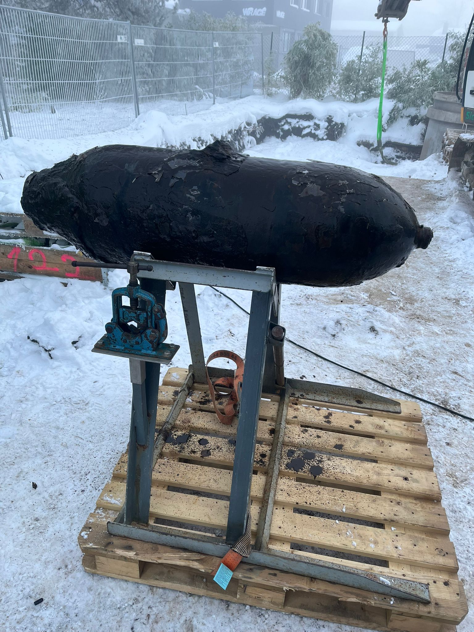 Eine entschärfte, stark verschmutzte 250-Kilogramm-Weltkriegsbombe liegt auf einer Metallhalterung, die auf einer Holzpalette im verschneiten Außenbereich steht.